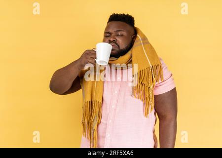 Traitement de la grippe. Portrait d'un homme malsain malade de la grippe portant une chemise rose enveloppée dans une écharpe chaude, buvant du thé chaud, traitant la grippe. Studio intérieur tourné isolé sur fond jaune. Banque D'Images