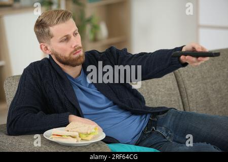 homme mangeant un sandwich et changeant de chaîne de télévision avec télécommande Banque D'Images