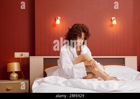 Une femme applique de la crème pour les pieds en étant assise dans un peignoir sur le lit Banque D'Images