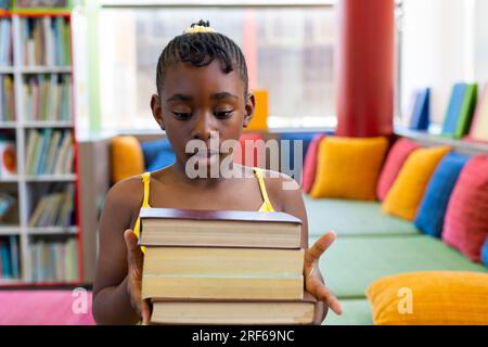 Écolière afro-américaine choquée tenant des livres au-dessus du canapé avec des oreillers colorés Banque D'Images