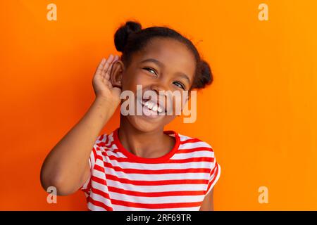 Écolière afro-américaine faisant geste d'écoute sur fond orange à l'école primaire Banque D'Images