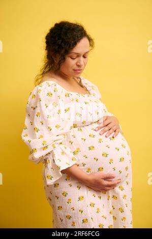 Femme enceinte, femelle gravide attendant un bébé, caressant doucement son gros ventre, appréciant les coups de pied de bébé, isolé sur fond de studio jaune. Pregna Banque D'Images