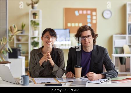 Portrait de vue de face de deux jeunes gens d'affaires homme et femme assis au bureau dans le bureau et souriant à la caméra ensemble Banque D'Images
