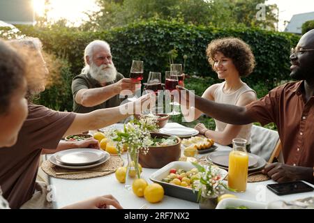 Heureux membres interculturels de la grande famille griller avec des verres de vin rouge tout en étant assis près de la table servie à l'extérieur et célébrant les vacances Banque D'Images