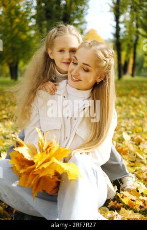 La famille s'amuse dans le parc. Mère est assise sur le sol avec un bouquet de feuilles d'automne, la fille embrasse sa mère par le cou, sourit et regarde t Banque D'Images