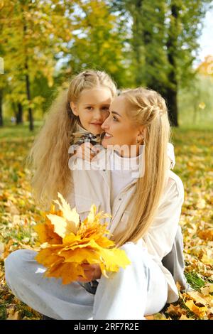 La famille s'amuse dans le parc. Mère est assise sur le sol avec un bouquet de feuilles d'automne, la fille embrasse sa mère par le cou, sourit et regarde t Banque D'Images