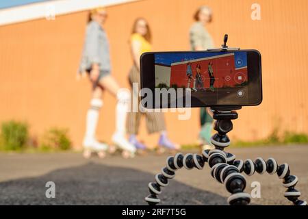 Concentrez-vous sur trépied avec smartphone lors du tournage vidéo de trois jeunes patineuses à roulettes féminines sympathiques en vêtements décontractés posant contre un mur brun Banque D'Images