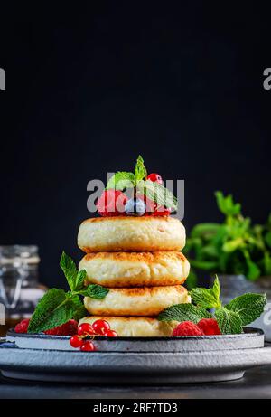 Crêpes épaisses au fromage cottage aux myrtilles, framboises et groseilles rouges, décorées de menthe fraîche sur une assiette grise, délicieux petit déjeuner. Tabl. Noir Banque D'Images