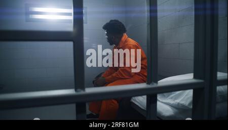 Détenus afro-américains en centre de détention ou en établissement correctionnel. Homme déprimé en uniforme orange assis sur le lit de prison et regarde la fenêtre barrée. Les prisonniers purgent une peine d'emprisonnement dans une cellule de prison. Banque D'Images