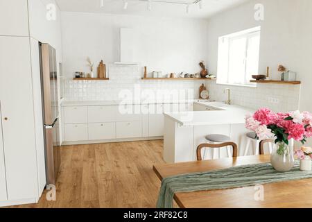 Intérieur de cuisine moderne. Belles pivoines dans le vase sur la table en bois et élégante cuisine blanche avec des appareils dans la nouvelle maison scandinave. Kitchenette minimale Banque D'Images