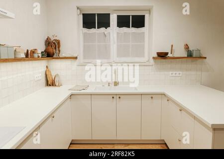 Intérieur de cuisine moderne. Armoires de cuisine blanches élégantes, robinet en laiton et évier en granit, étagères en bois avec ustensiles et lumière du soir dans nouveau scandin Banque D'Images