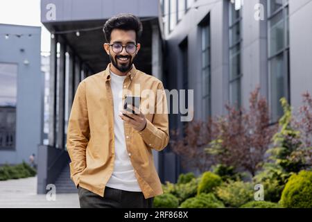 Jeune homme d'affaires prospère marchant dans la ville en dehors de l'immeuble de bureaux, hispanique souriant heureux avec les résultats de réalisation, tenant le téléphone, en utilisant l'application en ligne à l'extérieur. Banque D'Images