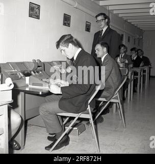 Années 1960, des étudiants de commerce masculins historiques et bien habillés dans une classe assis à des bureaux près du mur, sous la supervision d'un jeune enseignant adulte, Middletown, USA. Dans l'image, un étudiant utilisant une perforatrice de carte d'impression IBM 26. À cette époque, la carte perforée était l'unité d'exploitation de base du système comptable IBM. Étudiants bien habillés, tous portant un col et des cravates. Jeune enseignant masculin également en costume et cravate. Banque D'Images