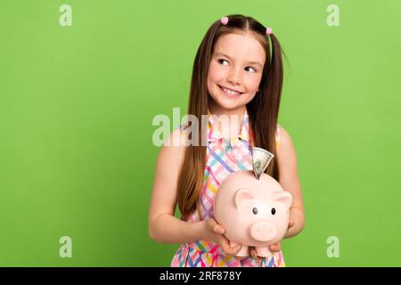 Photo de bras d'écolier positifs mignons tiennent le cochon de banque d'argent regarder l'espace vide isolé sur fond de couleur verte Banque D'Images