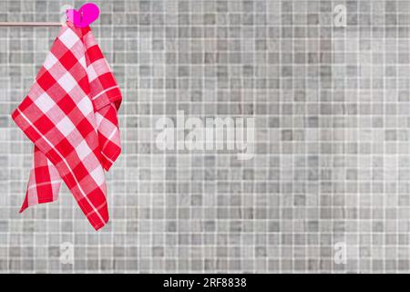 Une serviette de cuisine rouge à carreaux accrochée sur une tringle à vêtements contre un mur de carreaux de céramique en mosaïque gris beige flou abstrait. Espace pour l'affichage de votre produit lun Banque D'Images