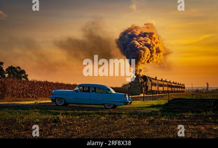 Une vue d'un train de passagers à vapeur antique approchant au lever du soleil avec une tête pleine de vapeur et de fumée voyageant à travers les terres agricoles avec une voiture antique attendant qu'il passe Banque D'Images