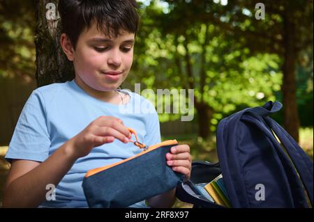 Caucasien beau garçon adolescent, élève intelligent de l'école primaire ouvrant son étui à crayons tout en faisant ses devoirs dans le parc après la classe. Retour à l'école le n Banque D'Images