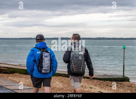 deux hommes portant des sacs à dos marchant le long d'une plage ensemble au bord de la mer par un jour couvert sur l'île de wight. Banque D'Images
