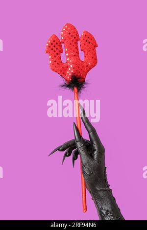 Main de sorcière avec fourche de diable rouge sur fond violet. Fête d'Halloween Banque D'Images