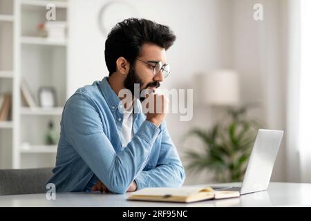Pensive Young Indian Male Freelancer dans les lunettes de travail avec ordinateur portable à la maison Banque D'Images