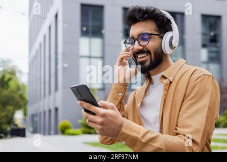 Jeune homme hispanique dans les écouteurs utilise une application pour écouter de la musique en ligne et des podcasts radio, un étudiant sourit heureux et tape au téléphone. Banque D'Images