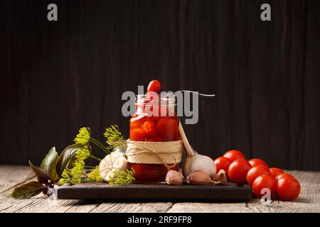 Tomates cerises marinées dans un bocal ouvert sur une planche en bois sombre, une tomate sur une fourchette vintage sur un bocal, fond sombre, gros plan. Banque D'Images