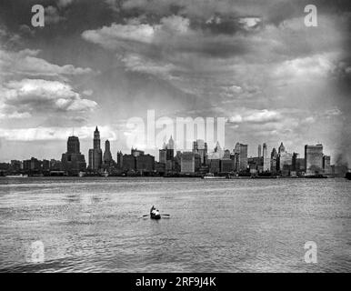New York, New York : c. 1927. Deux hommes dans une barque avec un drapeau américain pour un Bowsprit partent du New Jersey à travers le fleuve Hudson vers Lower Manhattan. Le Woolworth Building est le plus haut à gauche, puis le plus haut à droite avec le dôme rond est le Singer Building, et le double Equitable Building est au sud de cela. Banque D'Images