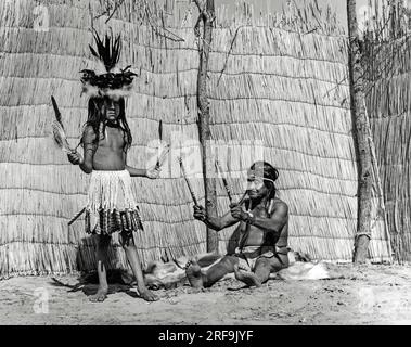 Californie : c. 1930 les hommes de médecine enseignaient leurs chants et danses à de jeunes garçons choisis pour perpétuer les traditions de la tribu. Ce medecin Yokut joue ses bâtons cassés en les secouant à temps jusqu'à sa chanson. Le costume de danse est une jupe en duvet d'aigle bordée de plumes de faucon à queue rouge. Le chapeau est un panier décoré de duvet d'aigle et de plumes de corbeau et de pie. Banque D'Images