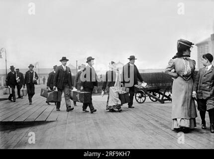 New York, New York : environ 1895 immigrants portant leurs bagages alors qu'ils arrivent à Ellis Island dans le port de NY. Banque D'Images