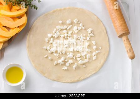 Roulez la pâte avec du fromage feta émietté, bol avec de l'huile d'olive, rouleau à pâtisserie, planche à découper avec de la citrouille tranchée et du thym frais sur fond blanc, t Banque D'Images