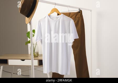 T-shirt blanc, chapeau et pantalon brun accroché à la tringle à vêtements avec espace de copie sur fond blanc Banque D'Images