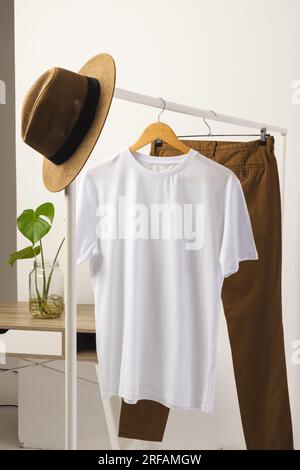 T-shirt blanc, chapeau et pantalon brun accroché à la tringle à vêtements avec espace de copie sur fond blanc Banque D'Images