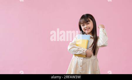 Une jeune fille asiatique joyeuse et mignonne tient ses livres et montre son poing serré tout en se tenant debout sur un fond de studio rose isolé. espace de copie Banque D'Images