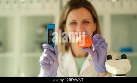 Femme scientifique tient dans les mains la mandarine et le testeur pour mesurer la quantité de nitrates dans le laboratoire chimique Banque D'Images