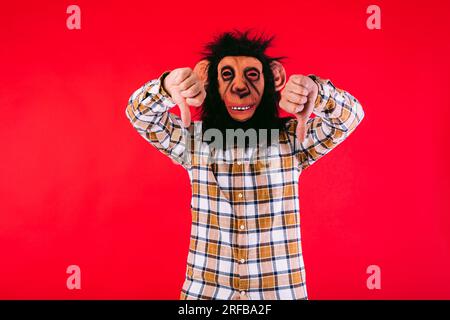 Homme avec masque de singe chimpanzé et chemise à carreaux, pouces vers le bas, sur fond rouge. Banque D'Images