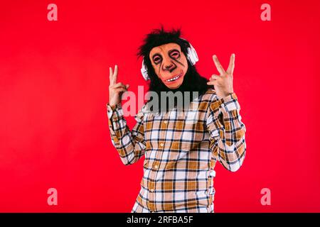 Homme avec masque de singe chimpanzé et chemise à carreaux, tenant ses doigts, écoutant son casque blanc, sur fond rouge. Banque D'Images