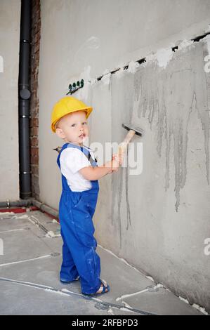 Toute la longueur de bébé garçon tenant le marteau et regardant la caméra tout en se tenant près du mur. Enfant portant un casque de sécurité et une combinaison de travail tout en utilisant l'outil de construction à la maison pendant la rénovation. Banque D'Images