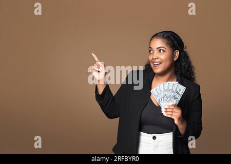 Heureuse dame latine plus taille tenant un ventilateur de dollar dans sa main et pointant de côté à l'espace libre, fond de studio brun Banque D'Images