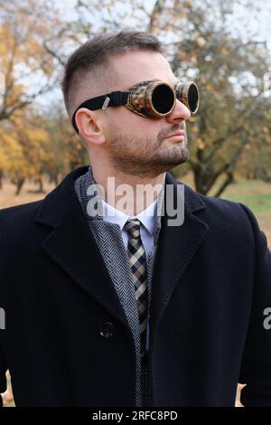 Un homme en costume d'affaires, manteau et lunettes steampunk pose dans un parc d'automne. Photo verticale Banque D'Images