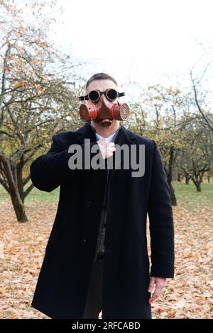 Homme en manteau noir, costume d'affaires et masque à gaz posant dans le parc d'automne. Photo verticale Banque D'Images