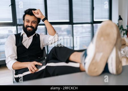 Portrait d'homme d'affaires barbu joyeux assis au bureau avec les pieds sur la table avec ordinateur portable, travail terminé, travail fait, terminé toutes les tâches Banque D'Images