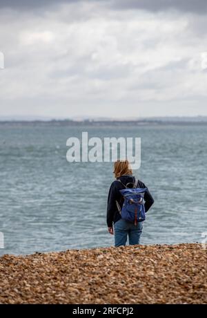 femme plus âgée debout sur une plage de galets regardant la mer. dame d'âge moyen portant un sac à dos debout seule sur une plage profondément dans la pensée. Banque D'Images