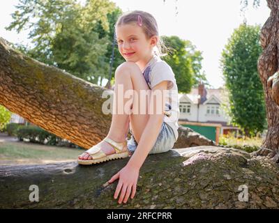 Portrait d'une jeune fille heureuse de huit ans assise sur une branche d'arbre dans le parc, Angleterre, Royaume-Uni Banque D'Images