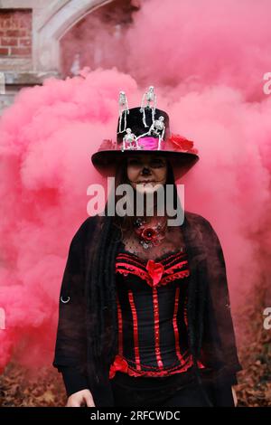 Une femme sous le couvert du baron Sabbath pose dans la fumée rouge. Le modèle est habillé d'un corset, d'un cardigan et d'un chapeau haut de gamme orné de figures de squelette. Banque D'Images
