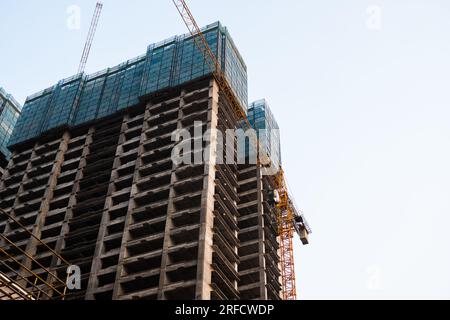 Grue de construction de tour jaune s'élève au-dessus d'un immeuble inachevé de plusieurs étages contre un ciel bleu clair. Construction monolithique moderne. Banque D'Images