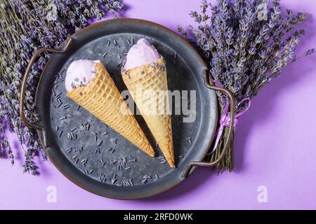 Deux glaces à la lavande en cônes gaufrés sur un plateau en étain et bouquets de lavande sèche sur fond lilas. Banque D'Images