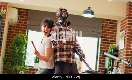 Couple diversifié aspirant des planchers en bois et montrant des mouvements de danse à la maison, s'amusant à faire des corvées. Joyeux homme et femme moderne dansant et chantant sur la musique, agissant drôle. Prise de vue à main levée. Banque D'Images