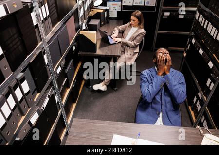 Vue de dessus du gestionnaire avec l'expression réfléchie, brainstorming des idées pour résoudre le rapport de bureaucratie dans la salle de stockage. Divers gens d'affaires travaillant des heures supplémentaires à la recherche administrative Banque D'Images