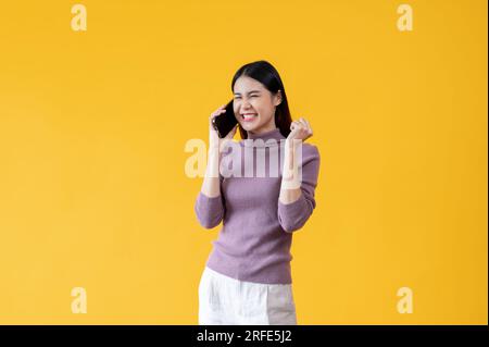 Une femme asiatique joyeuse et souriante parle au téléphone avec quelqu'un, se sentant heureuse et excitée après avoir reçu de bonnes nouvelles, se tenant contre un isol Banque D'Images