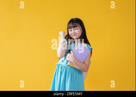 Une jeune fille asiatique joyeuse et positive dans une jolie robe bleue tient ses livres et montre son poing serré tout en se tenant contre un isolé Banque D'Images